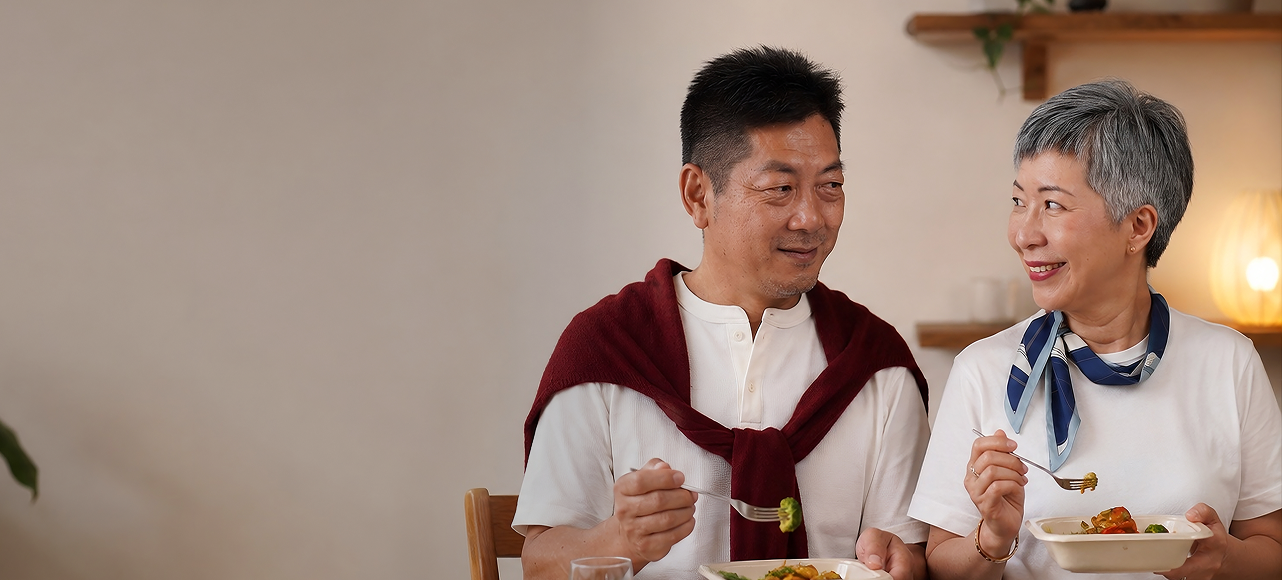 Two asian people sitting together, at a dining table, with a neutral background.